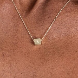 Op de afbeelding: Een gouden ketting met een klein, vierkant hanger met de letter "S" erop gegraveerd.