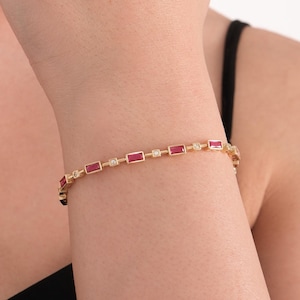 May include: A gold bracelet with alternating rectangular red and small round clear stones. The bracelet is displayed on a wrist, highlighting the jewellery's design and colour contrast.