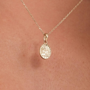 Puede incluir: Un collar dorado con un pequeño colgante ovalado. El colgante presenta un diseño detallado, posiblemente una figura o símbolo religioso. La cadena es delicada y el collar se muestra sobre un fondo color piel.