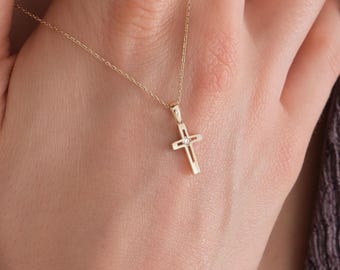 May include: A delicate gold cross pendant necklace. The cross is small and detailed, with a simple design. The necklace is shown against a neutral background, highlighting the jewelry's elegant simplicity. The chain is thin and appears to be gold.