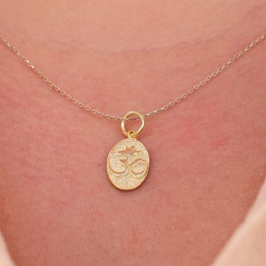 May include: A gold necklace with an oval pendant bearing the Om symbol. The pendant is suspended from a delicate chain. The necklace is displayed against a light-coloured shirt.