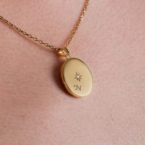 May include: A gold-toned oval locket pendant suspended on a delicate matching chain. The locket features a small, engraved starburst design with a central inset stone and the letter N engraved below it, creating a polished and refined accessory.