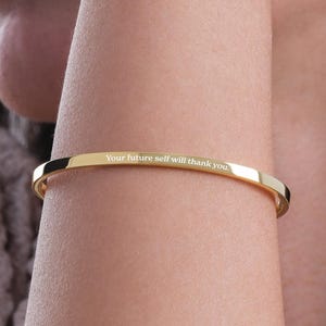 Könnte beinhalten: Ein polierter Armreif aus goldfarbenem Metall mit einem glatten, schmalen Banddesign. Die Außenfläche des Armreifs ist mit dem Text "Your future self will thank you." in einer klaren, gut lesbaren, weißen serifenlosen Schrift graviert.