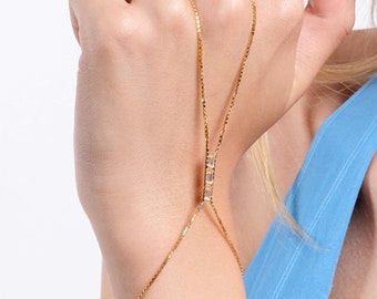 May include: A gold-toned hand chain featuring a delicate box chain design that connects a wrist bracelet to a central finger loop. The piece is accented with three small, clear, rectangular-cut stones positioned vertically along the center chain.