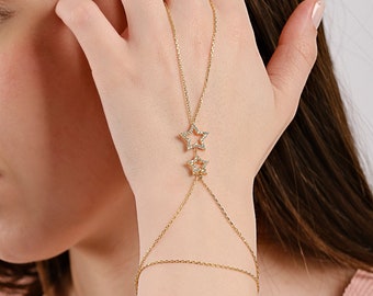 May include: A gold-toned hand chain bracelet featuring two delicate star-shaped charms encrusted with small clear stones. The jewelry piece connects a thin chain around the wrist to a central chain that extends upward to a loop for the middle finger.