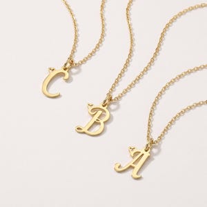 Può includere: Tre collane color oro con ciondoli a forma di lettere corsive C, B e A. Ogni ciondolo è sospeso a una catenina sottile e presenta un piccolo dettaglio a cuore integrato nella parte superiore della lettera, con finitura lucida.