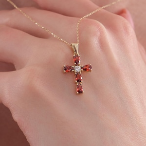 May include: A gold chain necklace featuring a cross pendant. The cross is composed of four pear-shaped red garnet gemstones arranged in a cross pattern, with a single small clear round gemstone set at the center where the arms intersect.