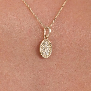 Può includere: Una collana con pendente ovale dorato. Il pendente presenta un rilievo dettagliato di una figura religiosa. La catena della collana è sottile e dorata. Il pendente è lungo circa 2 cm.