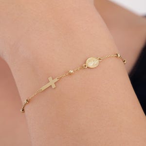 May include: A gold-colored bracelet featuring a cross charm and a circular charm with a religious figure. The bracelet has small, round beads and is worn on a wrist. The jewelry is made of a shiny, metallic material.