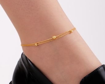 May include: A delicate gold-toned double-strand anklet featuring small, evenly spaced spherical beads along the chains. The jewelry piece is worn around the ankle, showcasing a fine link design that creates a layered appearance with a polished finish.