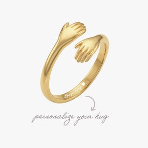 May include: A gold-toned open ring featuring two hands meeting in a hug design. The inner band is engraved with the text Love you always followed by a small heart symbol. Below the ring, the text personalize your hug is written in script.