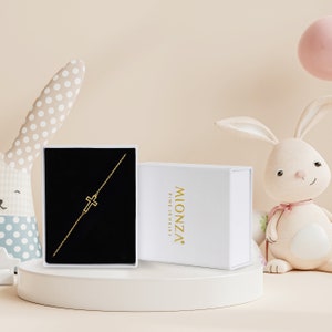A gold cross bracelet presented in a black box , accompanied by a decorative bunny figure.