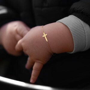 Baby's hand with a gold cross bracelet against a black stroller backdrop.