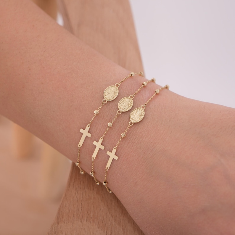 Four 14K solid gold rosary bracelets for women stacked on a wrist, each featuring a cross and Saint medal.