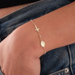 May include: A gold-colored bracelet featuring a cross and an oval pendant with a religious figure. The bracelet is delicate, with small beads and a thin chain. The bracelet is worn on a wrist, against a background of denim.