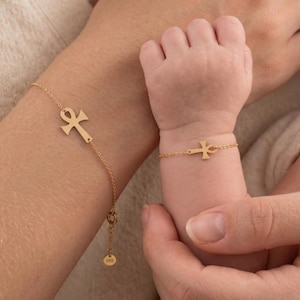 May include: Two gold bracelets featuring an ankh symbol charm. The larger bracelet has a delicate chain with a circular tag stamped with 585, while the smaller bracelet features a matching ankh charm on a thin gold chain, both worn on wrists.