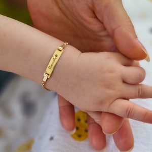 Close-up of a baby's wrist wearing a gold ID bracelet with the name 'Hannah' and a four-leaf clover symbol, held by an adult hand.