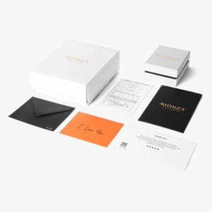 May include: A white jewelry box with the MIONZA logo, a smaller white box, a black envelope, an orange card with the text I Love You, a white jewel certificate, a black card with gold MIONZA text, and a white thank you card with a QR code.