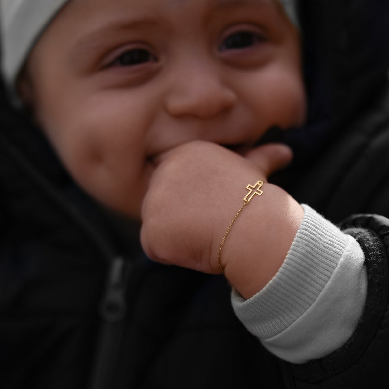 Smiling baby with a gold cross bracelet on the wrist, wearing a black jacket and a white beanie.