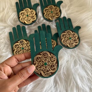 May include: A collection of dark green wooden Hamsa hand ornaments, each featuring a circular cutout with intricate gold-toned Arabic calligraphy arranged in a floral pattern and a small, centred green rhinestone accent.
