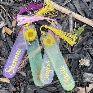 May include: Four resin bookmarks with gold lettering and colorful tassels. Each bookmark features a different name: Sienna, Taylor, and Olivia. The bookmarks are decorated with dried flowers and gold flakes, set against a purple and green background.