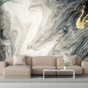 Puede incluir: Un salón moderno con un sofá seccional beige, una mesa de centro blanca y una alfombra gris. El punto focal es un gran mural de pared con un diseño de mármol en tonos grises, blancos y dorados. Una lámpara de pie y plantas completan la decoración.
