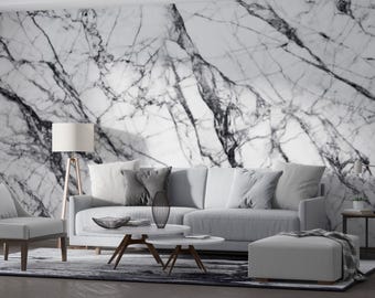 Puede incluir: Un salón moderno con una pared con diseño de mármol. La habitación cuenta con un sofá gris claro, un sillón a juego y una otomana blanca. Una lámpara de pie de madera y una mesa de centro completan la decoración. La habitación está decorada con cojines grises y una alfombra estampada.