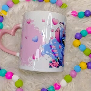 K&ouml;nnte beinhalten: Eine wei&szlig;e Keramiktasse mit einem rosafarbenen herzf&ouml;rmigen Henkel. Die Tasse zeigt eine blaue und rosa Comicfigur mit einer rosa Schleife und einem Zuckerstangen. Der Hintergrund ist ein rosa und blaues Aquarelldesign mit wei&szlig;en Herzen.