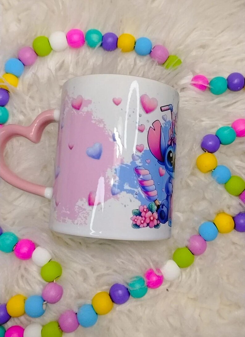K&ouml;nnte beinhalten: Eine wei&szlig;e Keramiktasse mit einem rosafarbenen herzf&ouml;rmigen Henkel. Die Tasse zeigt eine blaue und rosa Comicfigur mit einer rosa Schleife und einem Zuckerstangen. Der Hintergrund ist ein rosa und blaues Aquarelldesign mit wei&szlig;en Herzen.