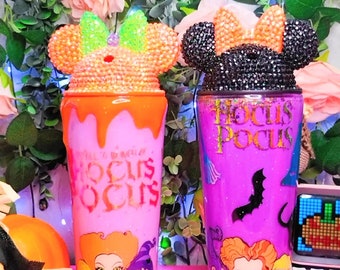 May include: Two pink and purple Halloween themed tumblers with glitter and a Mickey Mouse ear lid. One tumbler has a Hocus Pocus design with the Sanderson sisters from the movie. The other tumbler has a black glitter Mickey Mouse ear lid and says "Hocus Pocus" in gold lettering.