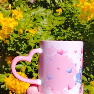 K&ouml;nnte beinhalten: Eine rosa Keramiktasse mit einem herzf&ouml;rmigen Henkel. Die Tasse hat einen rosa und wei&szlig;en Aquarellhintergrund mit verstreuten blauen und rosa Herzen.