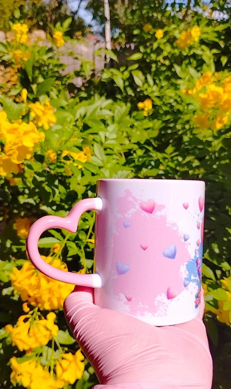 K&ouml;nnte beinhalten: Eine rosa Keramiktasse mit einem herzf&ouml;rmigen Henkel. Die Tasse hat einen rosa und wei&szlig;en Aquarellhintergrund mit verstreuten blauen und rosa Herzen.