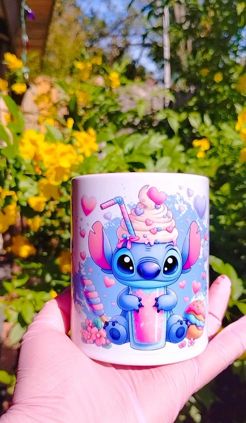 K&ouml;nnte beinhalten: Eine wei&szlig;e Keramiktasse mit einer blauen Comicfigur, die einen rosa Milchshake h&auml;lt. Die Figur hat gro&szlig;e Augen und eine rosa Schleife. Die Tasse ist mit rosa Herzen und einem wei&szlig;en Wirbelmuster verziert.