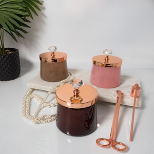 Puede incluir: Tres tarros de velas de vidrio con tapas de oro rosa y pomos de cristal. Los tarros están llenos de cera marrón, rosa y marrón oscuro. También se muestra un recortador de mecha de vela de oro rosa y un apagador de oro rosa.