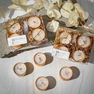 May include: Two sets of four scented tea light candles in clear plastic boxes with gold trim. The candles are in a brown paper filler. The boxes have labels with the names "Dreamy Cashmere" and "Allure".  There are also four individual tea light candles on a white surface.