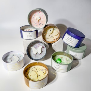 May include: Assortment of scented candles in glass jars with various decorative elements, including butterflies, flowers, and glitter. The candles are labeled with different scents, such as "Saffron Jasmine", "Bergamot Sandalwood", and "Pear Pomegranate".