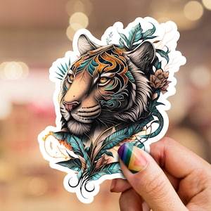 Puede incluir: Una pegatina con una ilustración detallada de la cara de un tigre, adornada con patrones turquesa y naranja y acentos florales. La pegatina tiene un borde blanco y está sujeta por una mano con una manicura colorida.