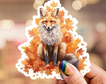 Op de afbeelding: Een rode vos met een witte borst en staart zit in een stapel oranje en gele herfstbladeren. De vos kijkt recht in de camera.