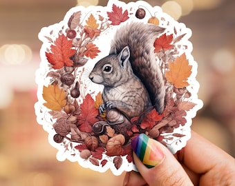 Puede incluir: Una pegatina con una ilustración detallada de una ardilla gris rodeada de hojas de otoño, bellotas y otros elementos del bosque. La ardilla sostiene una nuez en sus patas y mira hacia la izquierda.