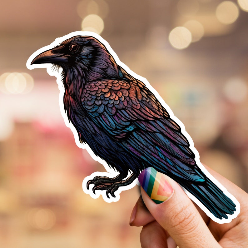 Op de afbeelding: Een sticker van een zwarte raaf met iriserende blauwe en paarse veren. De raaf kijkt naar links en heeft zijn vleugels uitgespreid. De sticker zit op de vinger van een persoon.