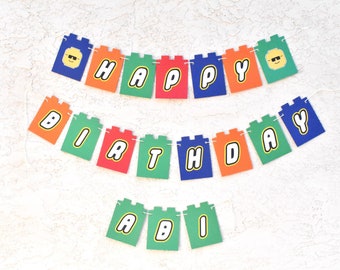 May include: A colorful birthday banner spelling "HAPPY BIRTHDAY ABI" in block letters. The banner is designed to resemble toy building blocks, with each letter on a different colored block. The banner includes a block with a smiling face wearing sunglasses.