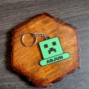 May include: A wooden keychain holder with a green and black Minecraft Creeper design. The keychain has the name "ARJUN" in white letters. The holder is a hexagon shape with a natural wood grain finish.