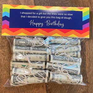 May include: A clear plastic bag filled with rolled up dollar bills tied with string. The bag is sitting on a wooden surface. A colorful card with the text "I shopped for a gift but the lines were so slow that I decided to give you this bag of dough. Happy Birthday" is behind the bag.