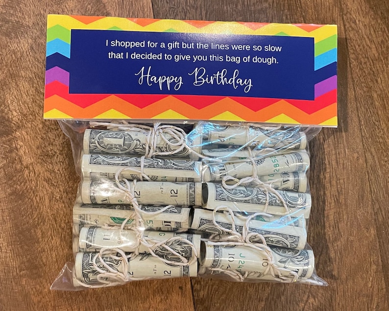 May include: A clear plastic bag filled with rolled up dollar bills tied with string. The bag is sitting on a wooden surface. A colorful card with the text "I shopped for a gift but the lines were so slow that I decided to give you this bag of dough. Happy Birthday" is behind the bag.