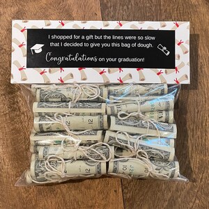May include: A white gift bag with a graduation theme. The bag has a printed message that says "I shopped for a gift but the lines were so slow that I decided to give you this bag of dough. Congratulations on your graduation!" The bag is filled with rolled up dollar bills.