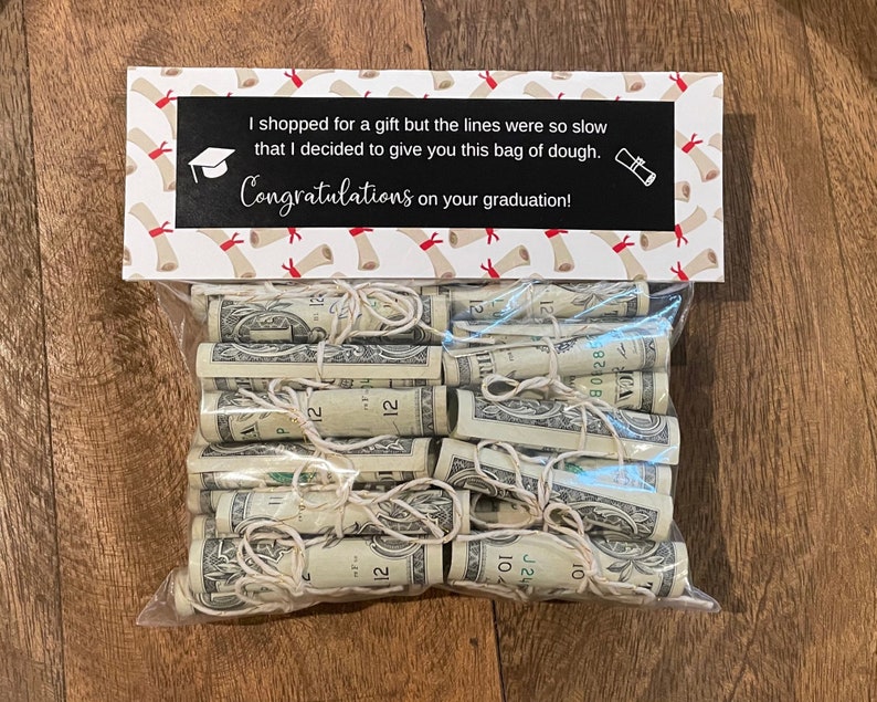 May include: A white gift bag with a graduation theme. The bag has a printed message that says "I shopped for a gift but the lines were so slow that I decided to give you this bag of dough. Congratulations on your graduation!" The bag is filled with rolled up dollar bills.