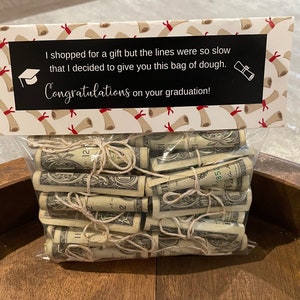 May include: A white gift bag with a graduation theme. The bag has a black label with the text "I shopped for a gift but the lines were so slow that I decided to give you this bag of dough. Congratulations on your graduation!" The bag is filled with rolled up dollar bills.
