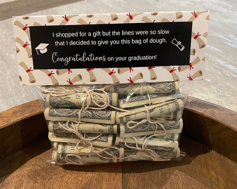May include: A white gift bag with a graduation theme. The bag has a black label with the text "I shopped for a gift but the lines were so slow that I decided to give you this bag of dough. Congratulations on your graduation!" The bag is filled with rolled up dollar bills.