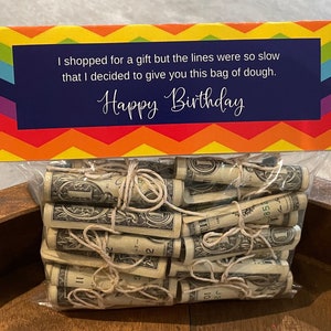 May include: A colorful birthday card with the text "I shopped for a gift but the lines were so slow that I decided to give you this bag of dough. Happy Birthday."  A clear plastic bag filled with rolled up dollar bills tied with twine.