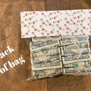 May include: A clear plastic bag filled with rolled up dollar bills tied with twine. The bag is sitting on a wooden surface. The bag is decorated with a white and red patterned paper with a design of red and white diplomas.